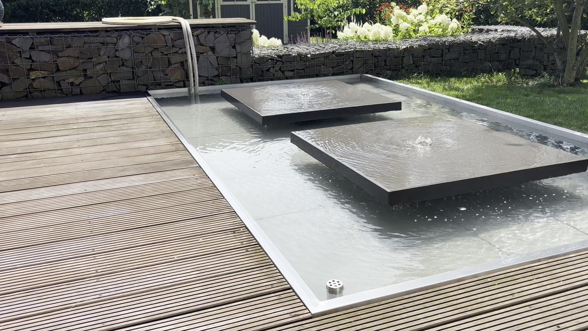Wasserbecken mit schwebenden Steinplatten neben einer Holzterrasse und Gabionenwand