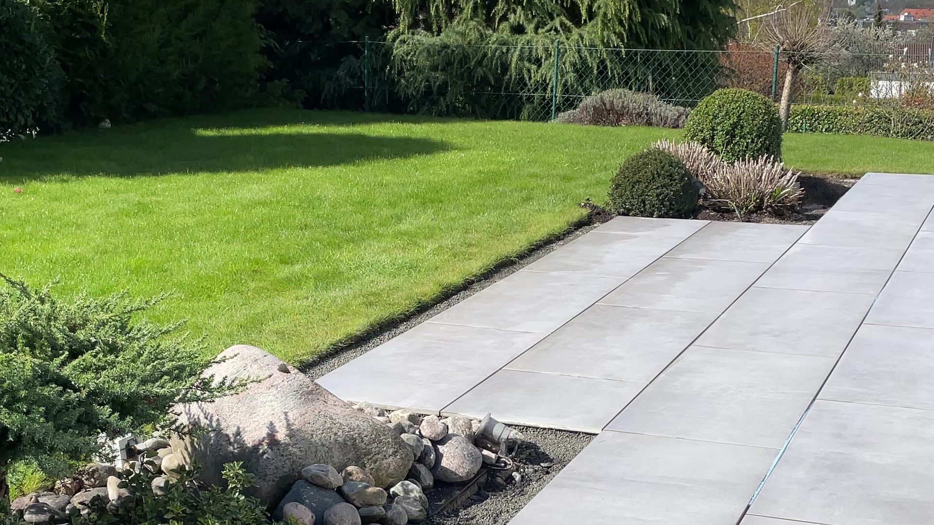 Terrasse aus großformatigen grauen Keramikplatten neben Rasen und Steinbeet